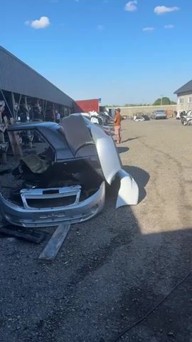 This media file depicts an outdoor junkyard filled with damaged and disassembled cars. The primary subjects are the vehicles themselves, ranging from car shells to individual parts like bumpers and car doors. Several people are visible, appearing to be workers or customers at the auto service and parts yard.

The scene is set in a gravel lot under a bright blue sky with scattered white clouds, suggesting it is daytime with pleasant weather. In the background, a building with a sign that reads "АВТОСЕРВИС" (AUTOSERVICE) is visible, indicating the location is a car repair and parts facility. The overall atmosphere appears practical and functional, typical of an auto salvage yard.

Notable details include the disarray of car parts and the presence of a red extension cord near one of the car wrecks. The sunlight casts shadows, further emphasizing the time of day. One of the visible car parts in the foreground has "PRIORA" and "LADA" written on it, identifying it as a part from a Lada Priora. The media appears to be a short, panning video showcasing the yard and its contents.