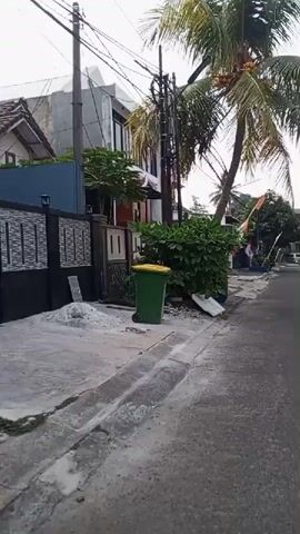 The video shows a street scene. In the foreground, there is a grey concrete sidewalk with a drainage channel. A green trash bin with a yellow lid is visible on the sidewalk. Behind the bin, there is a dense green bush and some construction debris. A black and grey fence runs along the edge of a property. To the right of the fence, a paved road stretches into the distance. A tall palm tree with sparse fronds is leaning over the road, with its upper branches partially obscuring the sky. Several small flags, possibly decorative or celebratory, are planted along the roadside. There are houses and other buildings visible on either side of the street. Some cars are parked along the road. The sky is bright and overcast. The video pans from left to right, slowly moving down the street.