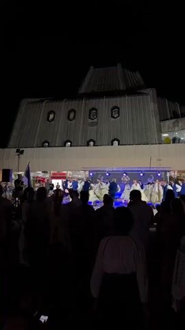 A night-time outdoor performance is underway on a raised stage in front of a large, angular building. A group of performers, dressed in white and blue traditional folk costumes, are dancing. A crowd of spectators, mostly silhouetted, watches from below the stage. The lighting is dim, with stage lights illuminating the performers and the building's facade. A flagpole with a blue and white flag is visible to the left of the stage.
