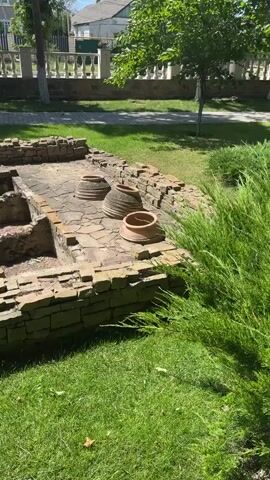 This video captures an outdoor scene of ancient ruins, possibly of a building or structure. The ruins are constructed from rough, tan-colored stones, forming low walls and tiered platforms or sunken areas. Several large, terracotta-colored pottery vessels are visible, some upright and others tilted on the stone floor of the ruins. The ruins are situated on a well-maintained grassy area, with green grass in the foreground and background. Beyond the ruins, a white picket fence lines a path, with trees and houses visible in the distance under a bright, sunny sky. The overall impression is one of historical preservation within a pleasant, contemporary environment.