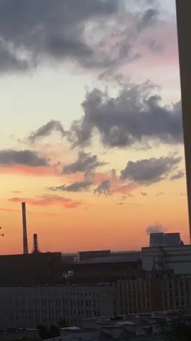 The video shows a cityscape at sunset. The sky is filled with dramatic clouds in shades of gray, pink, and orange. In the foreground, there are several buildings, including a tall smokestack and construction cranes. The sun is setting behind the buildings, casting a warm glow over the scene.