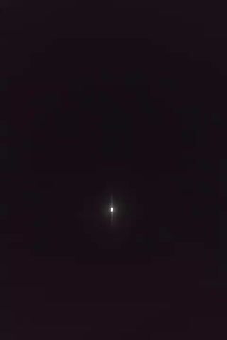 The video shows a celestial object, likely the moon, moving across a dark sky. The object appears bright and is visible as a white orb with some diffraction spikes, indicating it is a light source. The background is uniformly dark, suggesting a nighttime scene with no other visible celestial bodies or terrestrial objects. The object's movement is captured over a short period, showing a slight shift in its position against the dark expanse.
