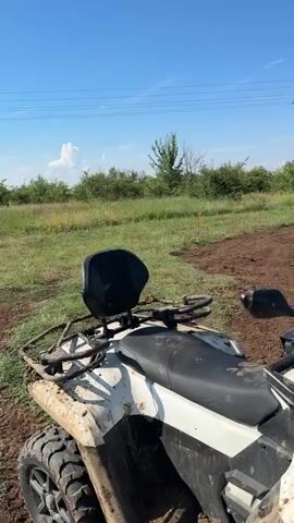 The video shows two ATVs in an outdoor environment. The first ATV is white and black and is in the foreground, partially obscuring the view of the second ATV. The second ATV is darker in color and is positioned further back in the frame. It appears to be a utility vehicle with a roof and a cargo bed. The ATVs are on a dirt or mud surface, with patches of grass and some vegetation visible in the background. There is a power line tower in the distance. The sky is blue with some clouds. The video is shot from a slightly elevated perspective, looking down on the ATVs. There are no people visible in the video. The scene appears to be a rural or off-road area.