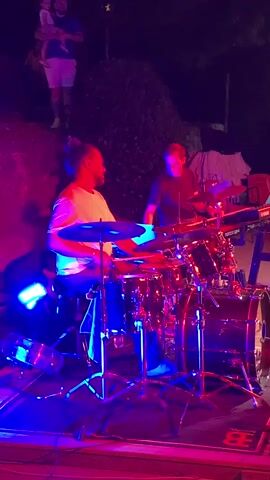 A man is playing a drum set on a stage. He is wearing a white t-shirt and dark shorts. Another man is standing behind him, playing a keyboard. He is wearing a black t-shirt. The stage is lit with blue and pink lights. In the background, there are trees and some people standing on a hill. One person is holding a baby. The stage floor has a logo on it.