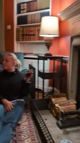 The video shows a group of people in a living room setting. Several people are sitting and standing in the room, holding glasses of wine. One person lifts another person's leg in the air while they are sitting on a couch. The living room has red walls, a patterned rug, and a bookshelf filled with books. There is a doorway in the background and a lamp on a table. The lighting in the room appears warm and inviting.