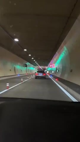 The video shows a car driving through a tunnel. The tunnel is brightly lit with lights of different colors, primarily green and red. There are barriers lining the road, and the speed limit is indicated as 80 km/h. Another car is visible in front. The perspective is from the driver's seat of the vehicle.