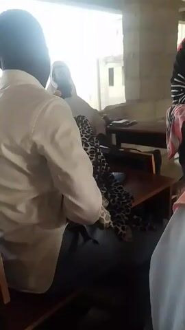 The video shows a woman in a striped black and white shirt and red headscarf talking in what appears to be a classroom setting with several other people.  A man in a white shirt sits at a desk holding a baby wrapped in a blanket with a leopard print pattern.  The classroom appears to be in a building under construction with exposed brick and an unfinished window. There are other desks and people partially visible in the shot.