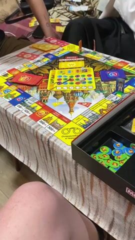 A group of people is playing a Russian version of the board game Monopoly indoors, likely in a home setting. The camera focuses on the colorful game board, which features Russian landmarks such as St. Basil's Cathedral at its center. Various property spaces, chance, and community chest cards are spread across the board. One person's hand rolls a white die onto the board, and then places a game token on a space. A game box, partially visible on the right, contains numerous green and blue circular game tokens. The table beneath the game board is covered with a striped, patterned tablecloth. Several players' hands and legs are visible around the table, indicating a social gathering. The game box has "MONOPOLE" printed on it, while the board clearly shows "МОНОПОЛИЯ" (Monopoly) in Russian.