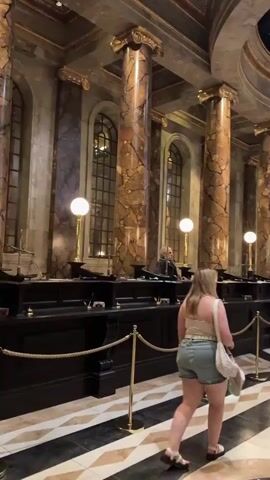 The video shows the interior of a grand hall, likely a bank or a place of high financial significance, characterized by tall, ornate marble columns and a dramatic, large, multi-tiered crystal chandelier hanging from the ceiling. The floor features a striking black and white geometric pattern. Several people are walking through the hall, mostly from right to left in the frame, in a queue or moving towards a row of counters. Behind the counters, individuals who appear to be staff members, dressed in formal attire, are interacting with customers or observing. The overall atmosphere suggests a bustling, formal environment with an emphasis on opulent design. One person, a woman with blonde hair wearing a white top and denim shorts, is in the foreground walking away from the camera, leading the viewer into the scene.
