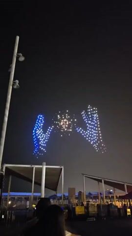 At night, a drone show displays a pair of hands, outlined in blue lights, reaching towards each other to form a circular shape in the center, also made of lights. Below, in the foreground, the silhouettes of people are visible, facing away from the camera, looking up at the sky. In the background, there are structures with lights, possibly part of a waterfront area or event venue. The overall environment appears to be an outdoor public space at night.
