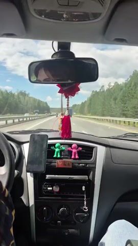 The video shows the interior of a car driving on a highway. In the foreground, a phone is mounted on the dashboard. Two small figures, one green and one pink, are also visible on the dashboard. A rearview mirror hangs from the windshield, reflecting the driver's face and the road ahead. A red tassel hangs from the rearview mirror. Outside the car, trees and a blue truck are visible on the side of the road. The sky is blue with some clouds. The overall impression is a person driving on a sunny day.