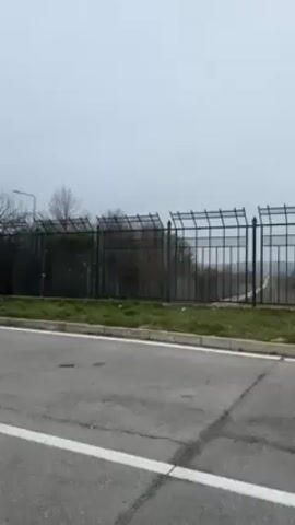 The camera pans across a green metal fence, revealing a long concrete wall in the distance. The wall is flanked by a dirt road and green fields. The sky is overcast.