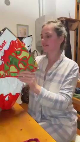 A young woman in a blue and white plaid pajama top sits at a wooden table, unwrapping a large Kinder chocolate egg. The egg is wrapped in red, white, and green foil. She tears open the foil with enthusiasm, pulling it back to reveal the chocolate egg underneath. The scene takes place in what appears to be a home setting with a cabinet and other furniture visible in the background.