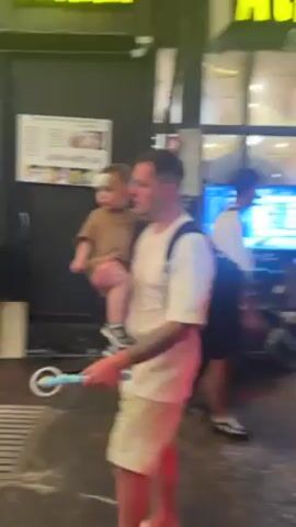 A man holds a child and is walking down a street at night. He has short hair, a white t-shirt and shorts, and tattoos on his arm. The child wears a brown outfit and a bandage on their head. The street is illuminated by string lights and neon signs of shops. Many people are walking on the street, and a scooter passes by. A woman records herself with a handheld camera.