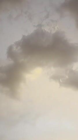 The video shows a sky filled with dramatic, dark clouds with some lighter patches of sky visible through them. As the video progresses, the sun begins to appear on the horizon, casting a bright, warm glow. The sun's rays are intense and seem to be breaking through the clouds. The lower portion of the video shows a body of water, likely the ocean, with gentle waves. The overall scene suggests a sunset or sunrise.