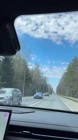 The video shows a view through the windshield of a car driving on a multi-lane highway. The sky is blue with scattered clouds. On either side of the road are trees. Several cars are visible on the road. There appears to be a display screen visible on the dashboard.