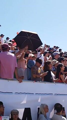 The video shows a crowd of people gathered around a body of water on a sunny day. Many people are standing behind a white barrier, some holding umbrellas for shade. In the water, there appears to be a race or competition taking place, with participants on paddle boards and in kayaks navigating a course marked by buoys and inflatable obstacles. A structure with scaffolding and Red Bull flags is visible in the background, suggesting a sponsored event. The sky is clear and blue.