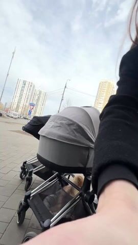 A woman wearing sunglasses and a black coat is standing outside on a paved walkway with a baby stroller in front of her. The stroller has a gray bassinet-style attachment on top and a small, fluffy dog in the storage area underneath. Behind the woman are tall apartment buildings and a cloudy sky. She adjusts the handle of the stroller and looks at the camera.