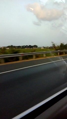 The video shows a view from inside a vehicle, likely a car or bus, looking out of a window. The perspective is horizontal, suggesting the video has been rotated. The dominant element is a dark asphalt road, possibly a highway, stretching into the distance. A white line marks the edge of the road. To the left of the road, there is a grassy verge with some scattered trees and what appears to be distant buildings or houses. The sky is overcast with some lighter patches that could be clouds or reflections on the window. The overall impression is of a roadside view during daytime, possibly a slightly gloomy or overcast day. There are no people or specific actions clearly visible within the frame.