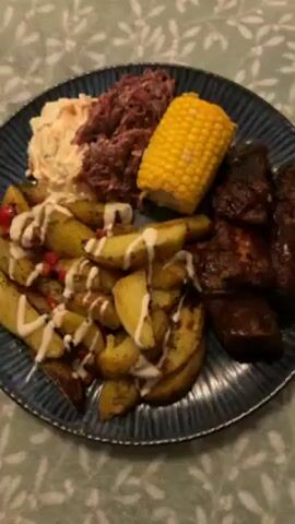 The video shows a plate of food that includes potato wedges drizzled with a white sauce, coleslaw, a pile of shredded red cabbage, a corn on the cob, and pieces of meat that look like ribs. The plate is placed on a surface with a light green and white floral pattern. A silver fork is placed near the plate. The camera moves around the plate slightly.