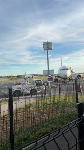 A towing vehicle is positioned beside a parked airplane on an airport tarmac under a blue sky with scattered clouds. Another airplane is visible in the background. The towing vehicle is white and has multiple wheels. The airplane in the foreground is white with yellow accents and appears to be a passenger jet. A black metal fence runs along the bottom of the frame, separating the viewer from the tarmac. The ground is a mixture of pavement and grass.