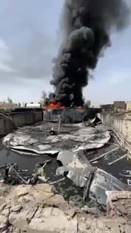 The video shows a large fire burning in what appears to be an industrial or storage facility. Thick black smoke billows upwards, obscuring the sky. The fire is centered on a circular structure, surrounded by debris and water. The surrounding area is also damaged, with crumbling brick walls and scattered metal fragments. There are no people visible in the video.