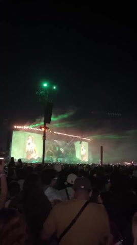 This video captures a vibrant night-time concert or music festival in Indio, United States of America. A large crowd of attendees fills the foreground and mid-ground, mostly standing and facing a brightly lit stage in the distance. The sky above is dark, contrasting with the dynamic light show.

On the main stage, two large screens are prominently displayed, each showing a close-up image of a female performer, likely a singer, with bright, warm lighting. Between these screens, a large, dark, elaborate structure, possibly resembling a dragon or monster, is visible, adding a dramatic element to the set design.

Green laser beams cut through the night sky, originating from various points around the stage and sweeping over the crowd. Smoke or fog hangs in the air, illuminated by the stage lights, creating an atmospheric effect. Multiple light fixtures and speakers are mounted on tall poles, further contributing to the visual spectacle. The crowd appears engaged, with many people holding up their phones, likely recording the performance or taking photos. The overall scene conveys an energetic and immersive concert experience. No discernible text is visible in the video.
