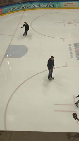The video shows an indoor ice rink with two people skating. One person wearing black clothing with long hair is skating dynamically. The other person is wearing a teal jacket and dark pants and is skating in a circle. There are red lines painted on the ice. Advertising appears above one of the rink's walls. There appears to be a camera tripod in the bottom right of the shot.