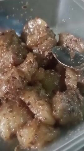 A close-up video shows a container of food, likely a dessert, cut into bite-sized pieces. The food appears to be coated in a sweet, granular substance, possibly sugar or a spice blend. A silver spoon is visible in the upper right corner of the frame, partially submerged in the food, suggesting it's being served or eaten. The lighting is dim, and the focus is soft, giving the food an appetizing, slightly blurred appearance.
