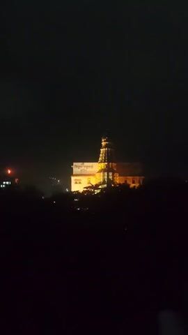 A temple is illuminated at night, with trees in the foreground obscuring the lower portion of the structure. A sign is visible on the temple, appearing to be in a language other than English. The sky is dark, suggesting it is nighttime.