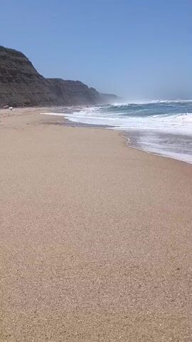 A stationary, eye-level shot of a sandy beach on a sunny day. The sand is light brown and appears dry. Waves are gently rolling in from the turquoise ocean, creating white foam on the shore. In the distance, a steep, rocky cliff face runs along the coastline under a clear blue sky. The cliff appears to be covered in sparse vegetation. There are a few small figures, possibly people, visible on the beach and along the base of the cliffs in the far distance, but they are too indistinct to identify. The overall impression is one of a remote and natural coastal landscape.
