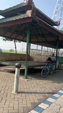 The video shows a man sitting on a bench inside a covered outdoor structure. The structure has a tiled roof with ornate eaves and is supported by green posts. In front of the structure, a blue bicycle is parked. The ground is paved with interlocking bricks. To the right of the structure, a broom and dustpan are visible. In the background, a field can be seen, with power lines and a large, white, lattice-like structure. The sky is clear and blue. The man is wearing a light-colored shirt and dark pants. He appears to be looking towards the right of the frame. The overall scene suggests a public outdoor space, possibly a park or rest area.