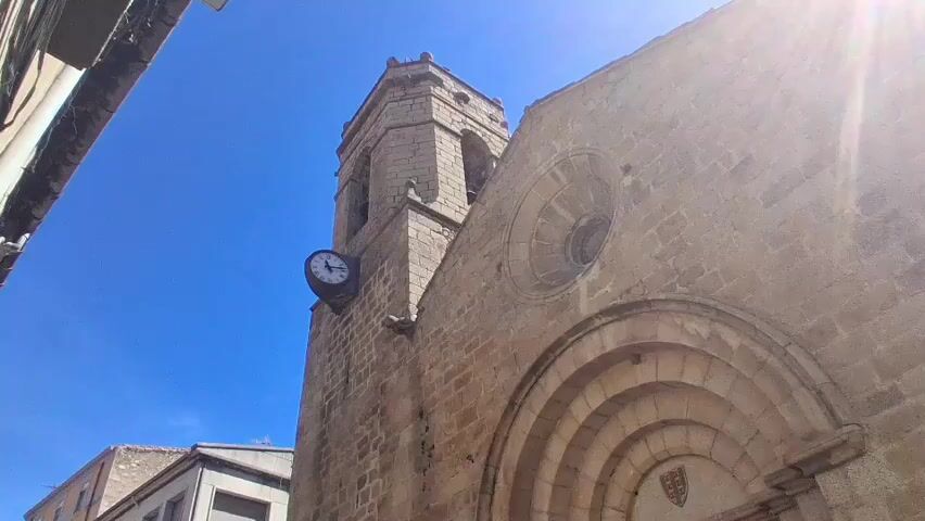 The camera pans down from the bell tower and clock of an old stone building, likely a church, to the front entrance with large wooden doors. A small green car drives in front of the building and a man is visible inside the car. The sky is bright blue and clear. The building appears to be located in a city or town with other buildings nearby.