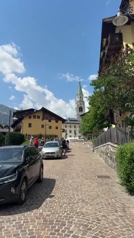 The video shows a charming, cobblestone street in Cortina d'Ampezzo, Italy. The scene is bathed in bright sunlight, suggesting a clear, pleasant day. The sky is a vibrant blue with scattered white clouds. Buildings with traditional alpine architecture line the street, featuring wooden balconies and warm-toned facades.

In the foreground, a black SUV is parked on the left. Further up the street, several cars are visible, including a silver hatchback and a dark-colored sedan. A few people are walking along the street, some appearing to be tourists, dressed in casual summer attire. They are scattered across the roadway and sidewalk.

Dominating the background is a striking white church tower with a green spire, a classic landmark of an Italian town. The presence of the mountains in the distance, though partially obscured, further reinforces the setting of Cortina d'Ampezzo, a renowned mountain resort town. The overall atmosphere is one of a relaxed, picturesque day in a European mountain village. There are no visible text elements.