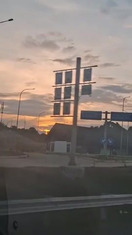 The video displays a time-lapse of a sunrise over a highway rest stop. The sky is filled with various shades of orange, pink, and yellow clouds, indicating the early morning hours. Several streetlights are visible, along with a building that appears to be a small rest area or booth. Road signs with directional arrows and text are also present. The view is from inside a moving vehicle, with the window framing the scene. No people are visible in the footage, only inanimate objects and the natural phenomenon of a sunrise.