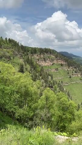 The video captures a panoramic view of a lush green mountain landscape with a valley below. Several people are present, some walking and others standing. One person is captured in the foreground, dressed in a pink t-shirt and black leggings, holding a phone and walking hand-in-hand with a man in a black jacket. Another woman in the background is wearing a white t-shirt and a plaid shirt, taking a photo with her phone. The sky is partly cloudy with dramatic white clouds against a blue sky. The scene is set on a grassy, rocky terrain.
