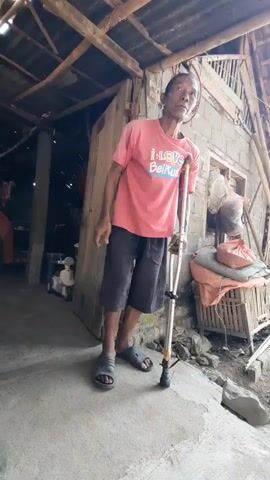 A full-body shot shows a man standing outside a rustic dwelling. He is wearing a pink t-shirt and dark shorts, and he is leaning on a forearm crutch. The man appears to be of middle age or older, with a thin build and a shaved head. His left arm is missing below the elbow. He is looking towards the camera. The background features a simple structure with a corrugated metal roof supported by wooden beams. There are various objects in the background, including what appears to be sacks of material, a fan, and possibly some household items. The ground is concrete, and there are some rocks and debris. The overall impression is one of a humble and possibly impoverished environment.