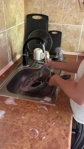 A woman is washing dishes in a kitchen sink. She is wearing a white tank top and dark striped pants. The sink is filled with soapy water, and she is scrubbing a pot with a sponge. There are dishes, cutting boards, and a faucet in the sink area. The kitchen has tiled walls and a wooden countertop.