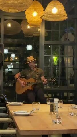 A man wearing a hat and olive green shirt plays an acoustic guitar and sings into a microphone. He is positioned behind a long, set dining table with plates, glasses, and salt and pepper shakers. The setting appears to be indoors, possibly a restaurant, with large paneled windows behind the musician. The ceiling has multiple decorative, textured light fixtures.