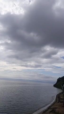 The video shows a vast expanse of the ocean under a cloudy sky. The water is a deep blue and appears calm with gentle ripples. The sky is filled with a mixture of dark, heavy clouds and lighter, scattered ones, with hints of sunlight breaking through in some areas. On the right side of the frame, a steep, vegetated cliff face meets the water, with a narrow strip of beach at its base. In the distance, the horizon stretches across the frame, with faint landmasses visible. A few small, dark objects, possibly buoys or distant boats, can be seen on the water. The overall atmosphere is moody and serene, with a sense of natural beauty and expanse.