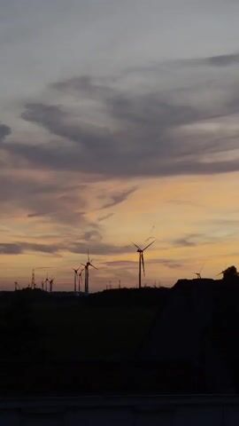 The video shows a silhouette of a wind farm against a twilight sky. The sky is a gradient of colors, with hues of purple, pink, and orange. Several wind turbines are visible in the distance, their blades either still or slowly rotating. The foreground is dark, with the silhouette of a building's roof and trees.