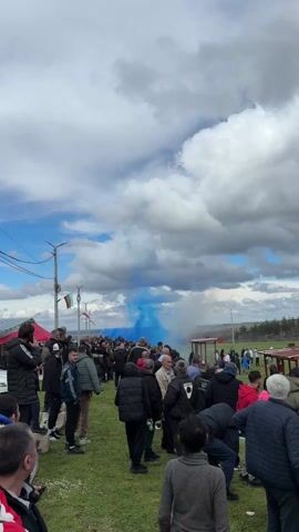 A wide shot captures a crowd of people gathered on a grassy area on a cloudy day. A plume of blue smoke rises behind the crowd. Most people are wearing jackets and hats. Some appear to be filming the smoke with their phones. The Bulgarian flag is seen on a pole to the left. A white plume of smoke is to the right of the blue plume. In the background, there is a soccer field and some covered stands. The camera appears to be hand-held and moves slightly. Someone speaks briefly at the beginning of the video.