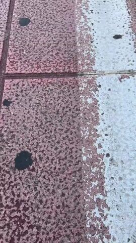 The video shows a low-angle view of a tiled sidewalk with a white line running down its center. The tiles are pink and have dark circular marks on them. The sidewalk appears to be next to a street with cars and buildings. A person on a bicycle is riding away from the camera down the sidewalk, following the white line. The scene suggests an urban environment, possibly a city street or promenade.