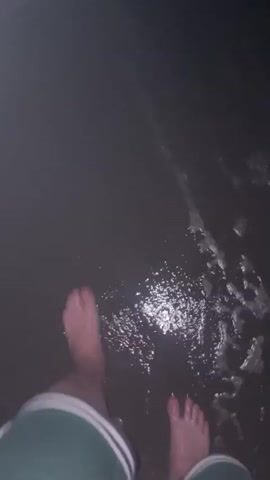 The video shows a first-person perspective of someone walking barefoot in shallow water at night. The water appears dark with some shimmering reflections, possibly from moonlight. Small waves and ripples are visible as the person moves their foot. The bottom is sandy or muddy. The person is wearing shorts with green and white stripes. The scene is dimly lit, suggesting it is dark outside.