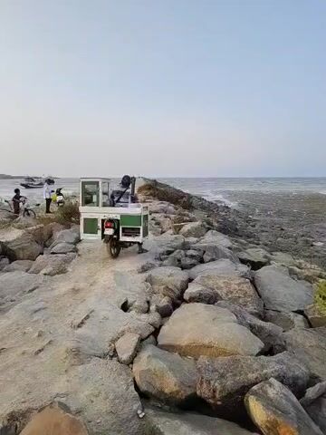 This is a ground-level, wide shot of a rocky shoreline on a clear day. The camera is facing a path leading away from the viewer and towards the ocean. The path is made of dirt and rocks, and on either side of the path are large, grey rocks. In the distance, the ocean stretches out to the horizon, with a lighter, possibly sandy or muddy area visible near the shore. There are a few boats anchored in the water in the far distance.

On the left side of the path, near the beginning of the frame, there are two people. One person is sitting on a bicycle, and another person is standing next to them, wearing a white shirt and dark pants. Further along the path, a person is seated on a motorbike that is pulling a small, green and white cart. The cart appears to have some items inside. The motorbike and cart are moving away from the camera, along the path. The overall atmosphere is calm and serene.