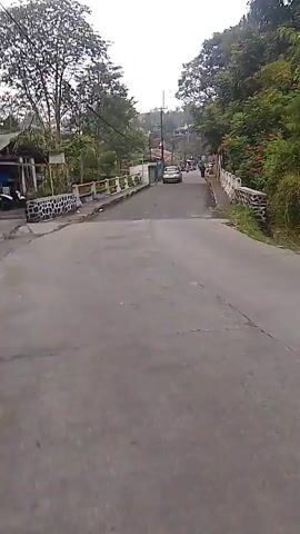 The video shows a paved road going uphill. The road is bordered by low walls and trees. There are a few vehicles and people on the road, and some buildings on the side. The scene appears to be a rural or suburban area.