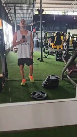 A man in a gym takes a selfie. He is wearing a white tank top with "I Heart Vietnam" printed on it, black shorts, and bright orange sneakers. He has a shaved head and a mustache, and has tattoos on his arms and face. The gym has mirrored walls, weightlifting equipment, and green artificial turf on the floor.
