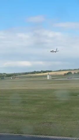 An airplane is in the process of landing. The plane is white with blue markings. It is flying in a clear blue sky with some scattered clouds. Below the plane is a green grassy field, and in the distance, there are trees and a runway. The camera is at ground level, looking up at the plane.