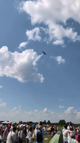 The video shows an aerial acrobatics performance. A small airplane is seen flying in a clear blue sky with scattered white clouds. The plane is leaving a white smoke trail as it executes a maneuver. Below the sky, a crowd of people is gathered, appearing to be watching the performance. Some people are holding up phones, likely to capture the event. The environment appears to be an open field with some trees and structures visible in the far distance. The perspective shifts slightly throughout the video, following the plane's movement across the sky.