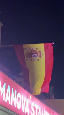 The video shows a Spanish flag hanging from a pole against a dark sky. Below the flag, a neon sign with pink letters reads "MANOVA STAMPING." The flag is red and yellow with a coat of arms on the yellow stripe. The flag appears to be gently waving.